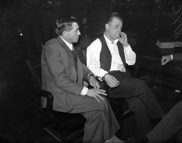 John Dillinger is handcuffed and guarded as he smokes during a court recess while Deputy Sheriff R.M. Pierce, left, looks on during Dillinger's hearing at Crown Point, Indiana in the first weeks of February 1934. Dillinger was charged with killing police officer William O'Malley, 43, during a bank robbery in East Chicago, Indiana. (Chicago Tribune historical photo)