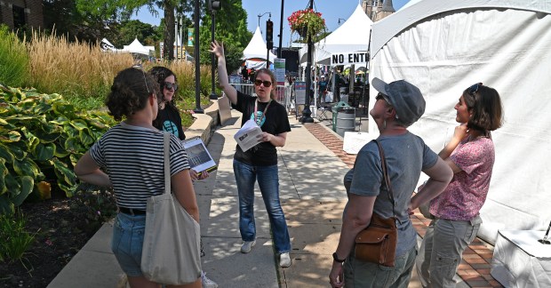 Arm raised, Museum Manager Emily England of Skokie and of the Skokie Heritage Museum is in front of Skokie Village Hall with the tour group on Oakton Street during the Historic Downtown Skokie Walking Tour on Aug. 25, 2024 in downtown Skokie. (Karie Angell Luc/Pioneer Press)