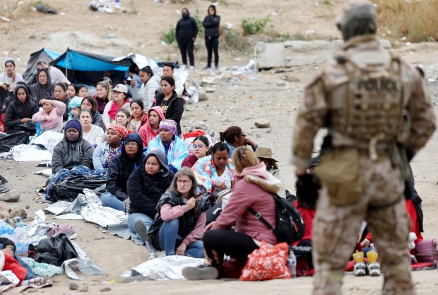 Immigrants seeking asylum in the U.S., who are stuck in a makeshift camp between the U.S.-Mexico border walls, sit as a U.S. Customs and Border Protection officer keeps watch while other migrants are lined up to be transported on May 13, 2023, in San Diego. Some of the migrants at the open-air camp have been waiting for days for a chance to plead for asylum while local volunteer groups provide food and other necessities. (Mario Tama/Getty)