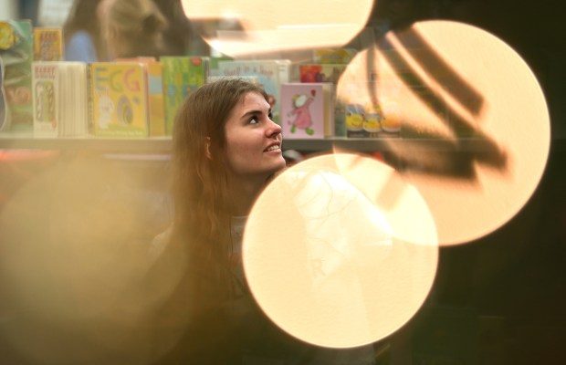 Through the view of an illuminated holiday tree, Alexandra Walocha, 17, of Barrington Hills, a junior at Barrington High School, is arranging children's books to offer to patrons in the school's library section at Barrington Giving Day's Winter Event on Dec. 14, 2024 at Station Middle School (215 Eastern Ave.) in Barrington. (Karie Angell Luc/Pioneer Press)