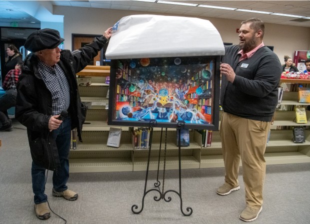 LaPorte artist Rocco Schiralli, left, unveils his original work "Beyond Eternal Blue Outer Space, Reading Can Transport You to Anyplace" with the help of Jesse Butz during the Winter Wonder Fest celebration at the Portage Public Library in Portage, Indiana, on Friday, Dec. 20, 2024. Butz is director of the Porter County Public Library System.(Andy Lavalley/for the Post-Tribune)