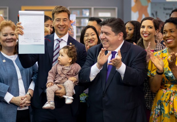 Illinois Secretary of State Alexi Giannoulias holds his 11-month-old daughter, Alexia, and the bill signed by Gov. JB Pritzker to prevent book bans on June 12, 2023, at the Harold Washington Library in Chicago, while Pritzker applauds. (Brian Cassella/Chicago Tribune)