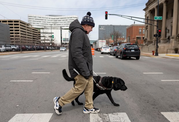 Peter Berg, who is blind, crosses South Ashland Avenue with his service dog, Lloyd, on March 18, 2025, in the Medical District. This intersection of South Ashland Avenue and West Polk Street does not have audible pedestrian signals and Berg relies on hearing the traffic stop to know when to cross. (Brian Cassella/Chicago Tribune)