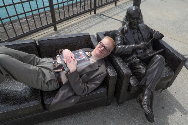 Author Walter Podrazik lies on the couch at the Bob Newhart sculpture on Navy Pier in Chicago with a copy of his book "Watching TV: American Television Season by Season," on April 29, 2025. (Terrence Antonio James/Chicago Tribune)